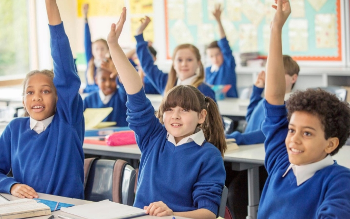 Children in classroom, hands raised as they engage with their learning