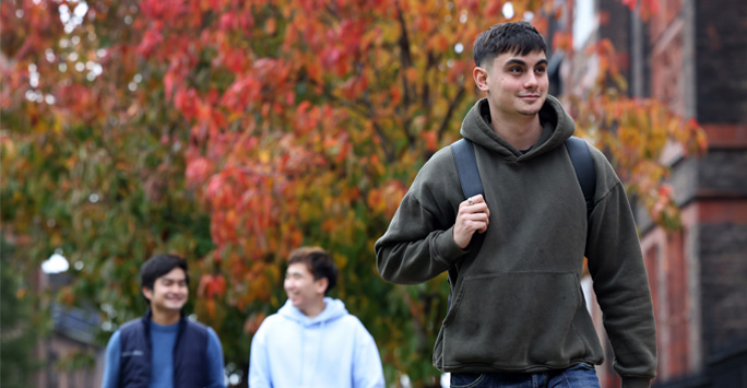 A student walks through campus. There are trees with orange leaves surrounding him.